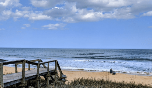 A serene beach scene with gentle waves, a wooden walkway, and a person sitting on the sand under a blue sky.