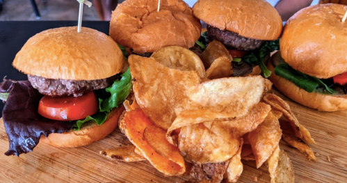 Three mini burgers with lettuce and tomato, served with a side of crispy potato chips on a wooden board.