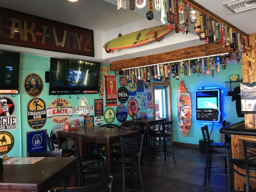 A vibrant bar interior with colorful beer signs, surfboards, and a jukebox, featuring wooden tables and chairs.