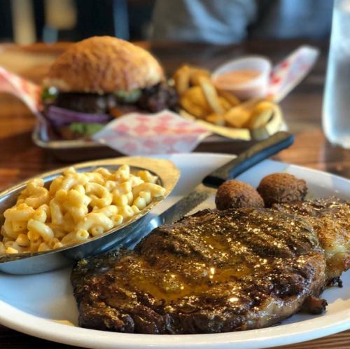 A plate featuring a juicy steak, macaroni and cheese, and fried sides, with a burger and fries in the background.