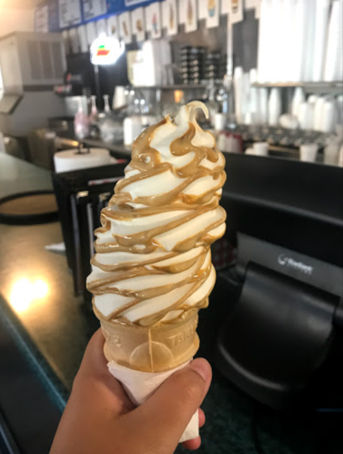 A hand holding a soft-serve ice cream cone topped with caramel drizzle, set against a café background.