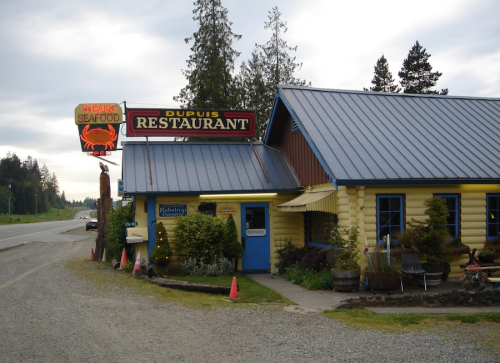A rustic seafood restaurant with a yellow exterior, blue roof, and a sign featuring a crab, surrounded by trees.