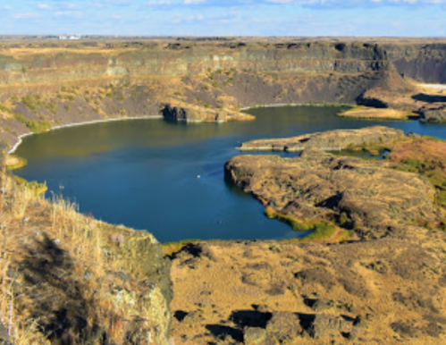 A scenic view of a canyon with a calm lake surrounded by rocky cliffs and sparse vegetation under a clear blue sky.