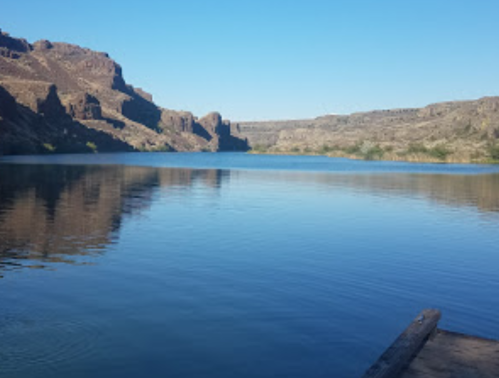 Serene lake surrounded by rocky cliffs under a clear blue sky, reflecting the landscape in calm waters.