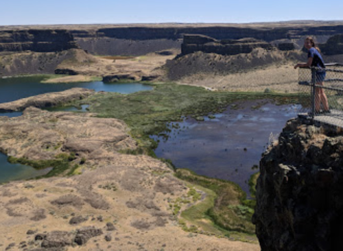 A person stands on a railing overlooking a vast canyon with lakes and greenery below.