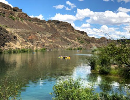 A serene river scene with kayakers, surrounded by rocky cliffs and lush greenery under a blue sky with fluffy clouds.