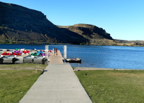 A scenic view of a dock with colorful boats, leading to a calm lake surrounded by hills under a clear blue sky.