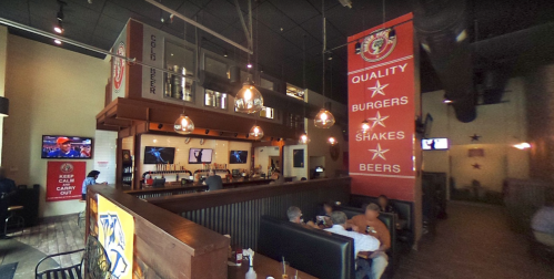 Interior of a casual restaurant featuring a bar, seating area, and banners promoting burgers, shakes, and beers.