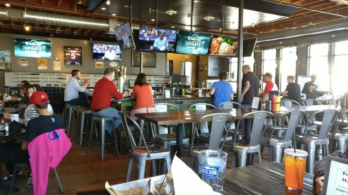 A lively restaurant interior with patrons at tables, watching sports on multiple screens, and enjoying food and drinks.