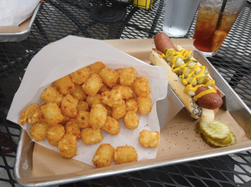 A plate with crispy tater tots and a hot dog topped with mustard, pickles, and onions on a tray.