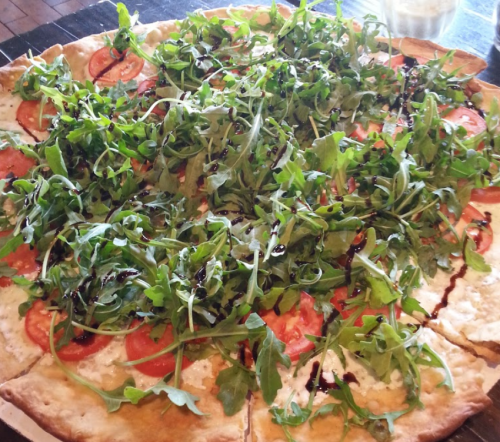 A large pizza topped with fresh tomatoes and a generous layer of arugula drizzled with balsamic glaze.