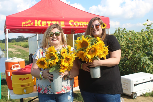 Two women hold large bouquets of sunflowers in front of a red tent labeled "Kettle Corn" on a sunny day.