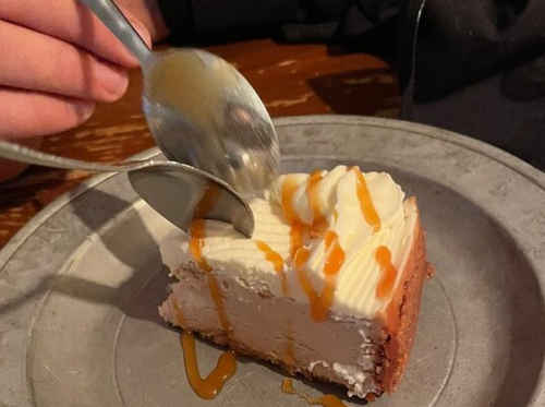 A hand holds two spoons above a slice of cheesecake drizzled with caramel sauce on a metal plate.