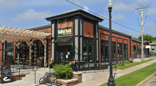 A modern restaurant with a wooden exterior, large windows, and outdoor seating area on a sunny day.