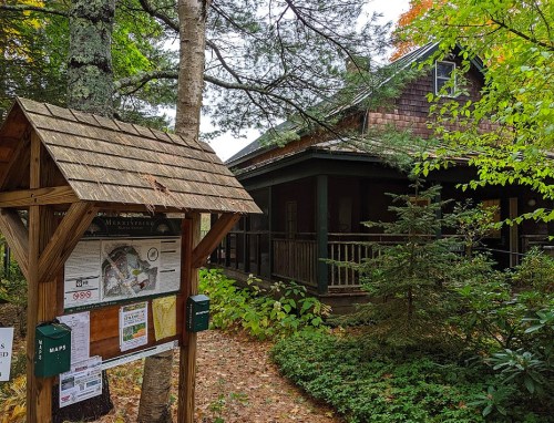 A wooden signpost with maps stands in front of a rustic cabin surrounded by trees and greenery.