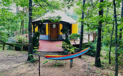 A colorful yurt surrounded by trees, with a hammock in the foreground and a wooden deck leading to the entrance.