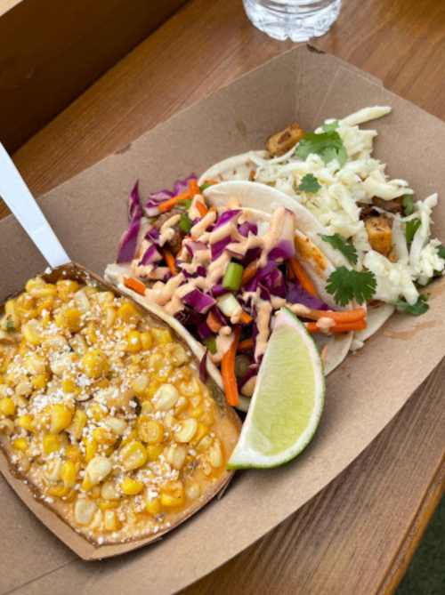 A paper tray with two tacos, one with slaw and the other with corn, topped with lime and drizzled sauce.