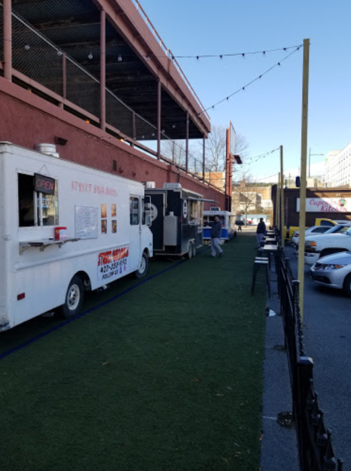 A food truck is parked beside a building, with people walking on a grassy area and cars nearby under a clear sky.
