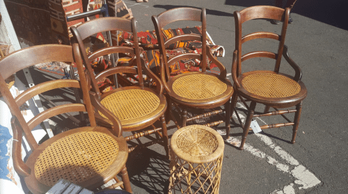 A row of wooden chairs with caned seats, alongside a small woven stool, set on a pavement.