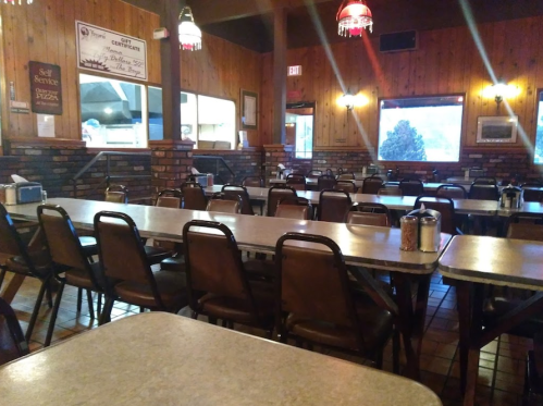 A spacious, rustic restaurant interior with empty tables and chairs, wooden walls, and large windows.