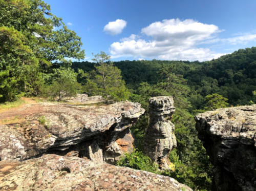 A rocky cliff overlooks a lush green valley under a blue sky with scattered clouds.