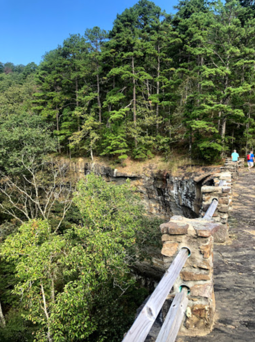 A stone railing overlooks a rocky cliff surrounded by lush green trees under a clear blue sky.
