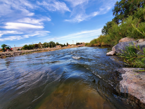 A serene river flows gently, bordered by rocks and greenery, under a blue sky with scattered clouds.