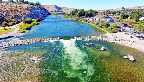 A scenic view of a river with a small waterfall, bridge, and lush greenery along the banks under a clear blue sky.