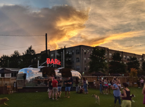 A lively outdoor scene at sunset with a food truck labeled "BARK" and people socializing, some with dogs.