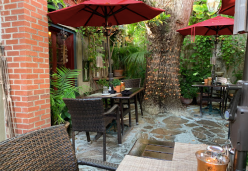 A cozy outdoor dining area with wicker tables, red umbrellas, and lush greenery surrounding a stone pathway.