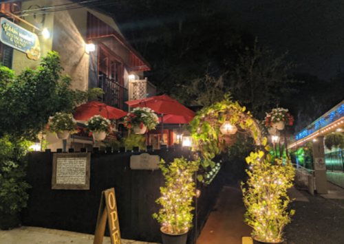A cozy restaurant entrance at night, featuring red umbrellas, greenery, and string lights illuminating the pathway.