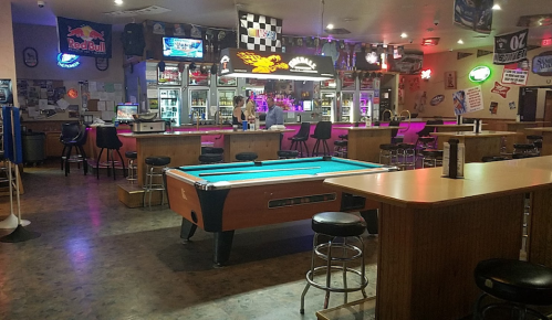 A dimly lit bar with a pool table, stools, and neon signs, featuring a bartender behind the counter.