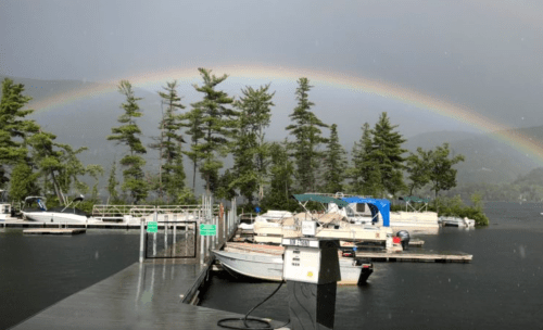 A rainbow arcs over a lake with boats docked and trees lining the shore, amidst a rainy atmosphere.