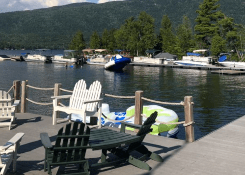 A peaceful lakeside scene with boats docked, green trees, and Adirondack chairs on a wooden pier.