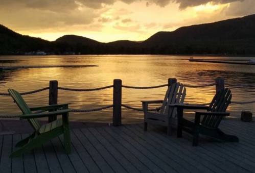 Two Adirondack chairs on a dock overlooking a calm lake at sunset, with mountains in the background.