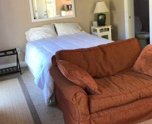 A cozy bedroom featuring a bed with white linens, a brown couch, and a lamp beside a small table.
