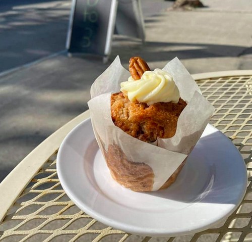 A freshly baked muffin topped with cream cheese frosting and a pecan, served on a white plate outdoors.