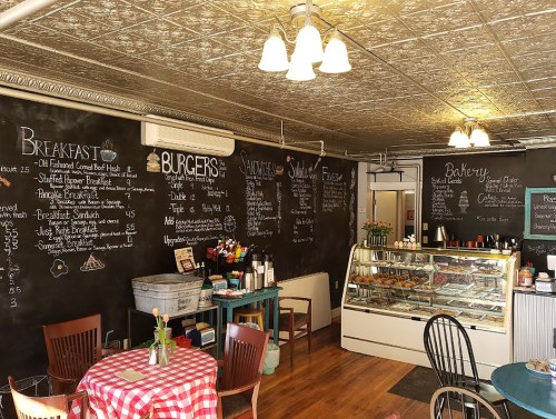 Cozy café interior with chalkboard menus, a display case of baked goods, and a checkered tablecloth dining area.