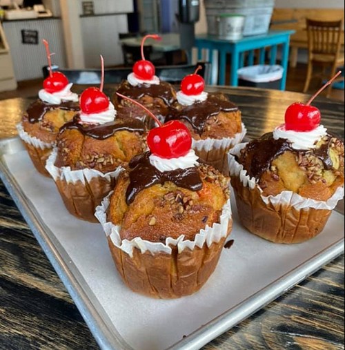 A tray of muffins topped with chocolate, whipped cream, and cherries, set on a wooden table.