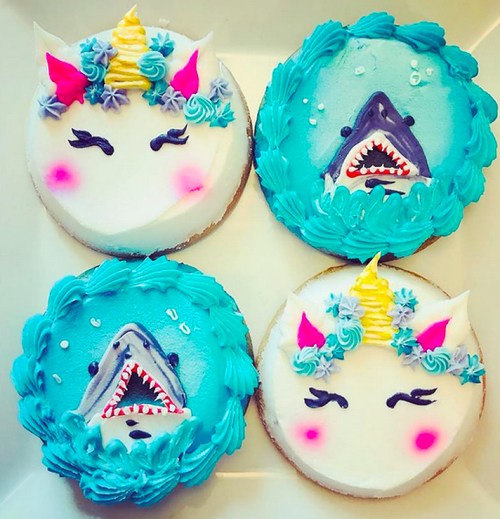 Four colorful cupcakes: two with unicorn designs and two with shark faces, all decorated with vibrant frosting.
