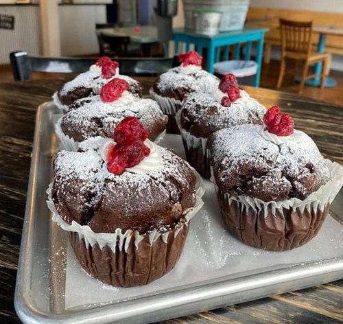 A tray of chocolate muffins topped with powdered sugar and raspberries, set on a wooden table.