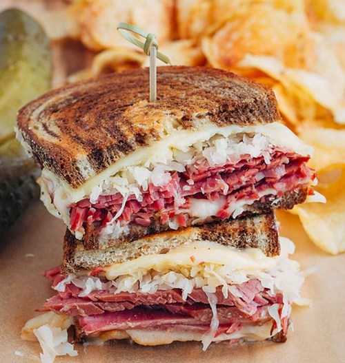 A stacked sandwich with layers of pastrami, sauerkraut, and melted cheese on toasted rye, garnished with a pickle.
