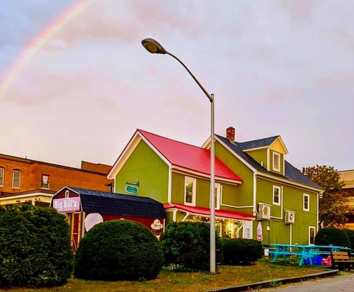 A colorful house with a red roof and green walls, under a rainbow, with bushes and a streetlamp in the foreground.