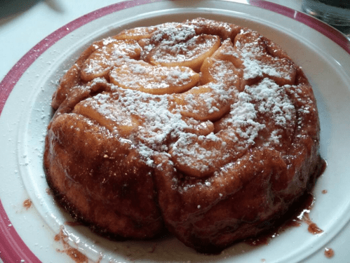 A round dessert topped with caramelized apples and dusted with powdered sugar on a white plate.