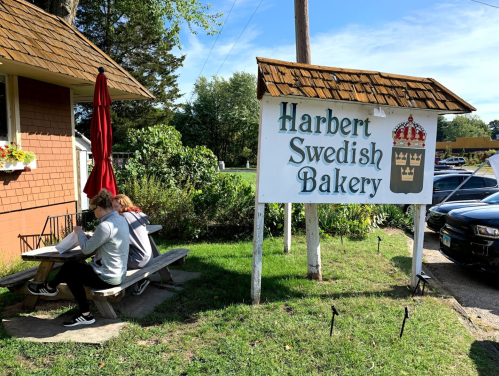 A sign for Harbert Swedish Bakery outside, with two people sitting at a picnic table nearby.