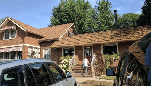 A charming house with a shingled roof, surrounded by greenery, and a child playing on the steps. Two cars parked nearby.