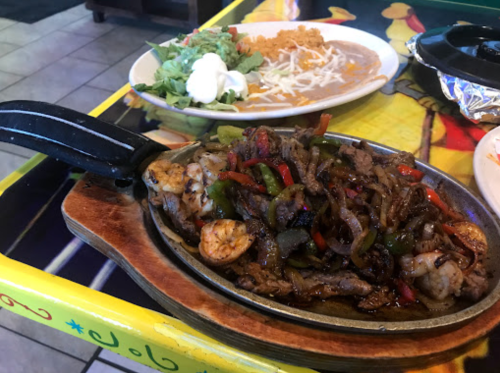 Sizzling fajitas with shrimp and vegetables on a skillet, accompanied by a salad and rice on a colorful table.