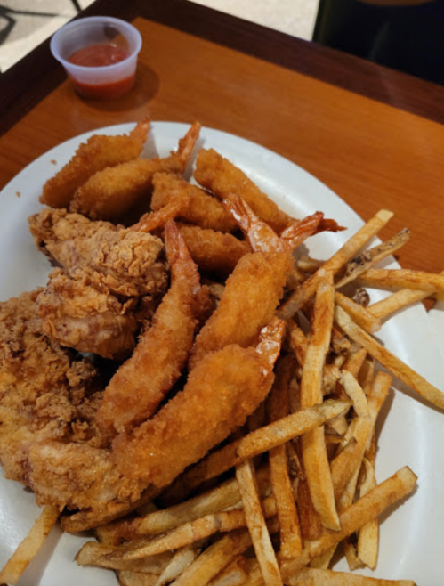 A plate of fried shrimp, crispy chicken, and golden French fries, accompanied by a small cup of dipping sauce.