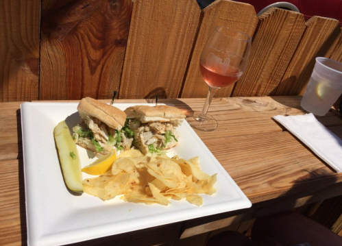 A plate with two small sandwiches, chips, a pickle, and a glass of rosé wine on a wooden table.