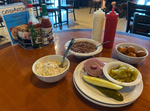 A table setting with bowls of coleslaw, baked beans, hushpuppies, pickles, and a menu for Catfish Kitchen.